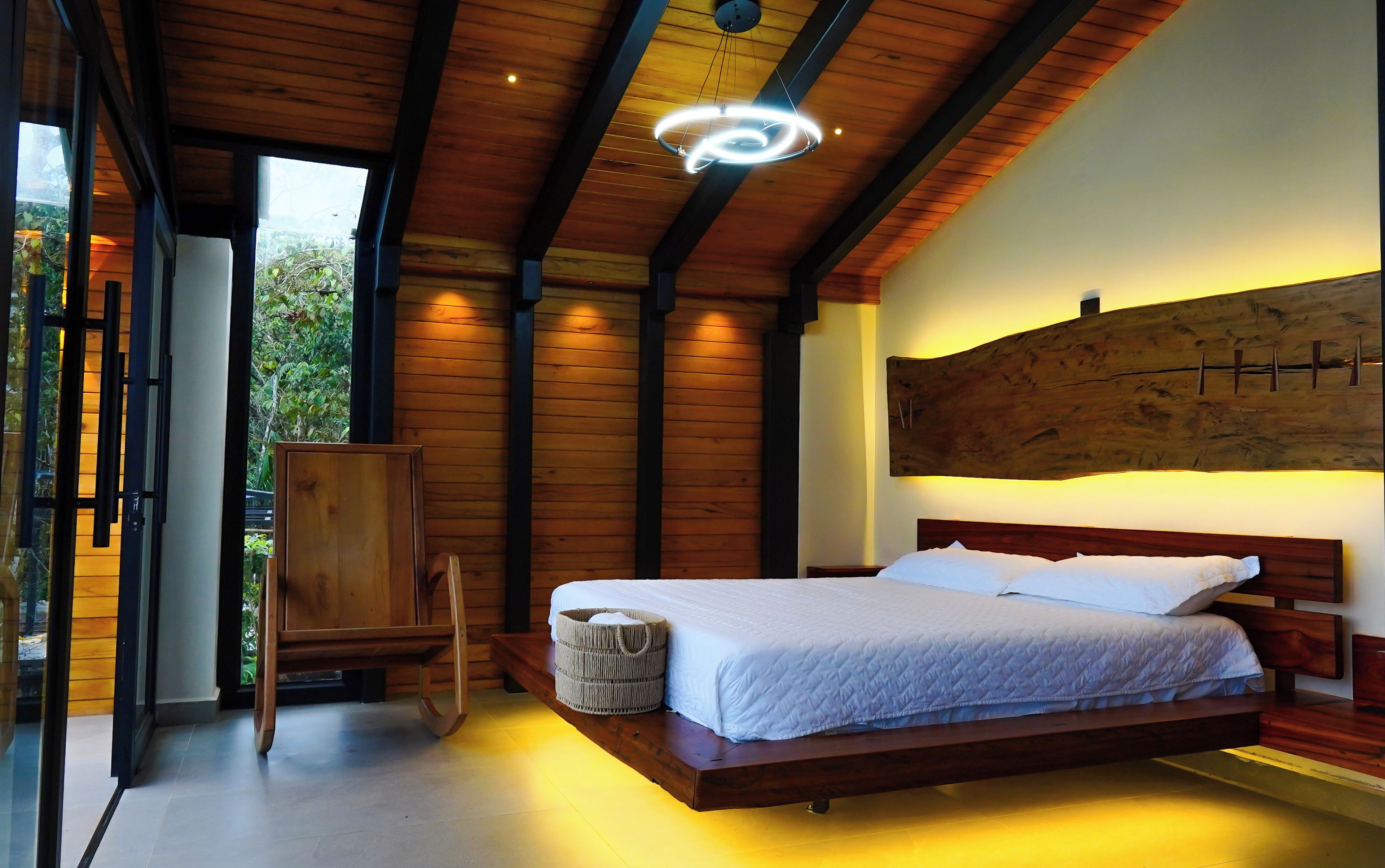 Un dormitorio con techo de madera, cama con ropa blanca, una gran obra de arte de madera en la pared, y vistas a través de un ventanal.