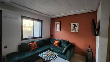 Ein Wohnzimmer mit einem grünen Sofa, einem Couchtisch, einem Fenster und einem an der Wand befestigten Fernseher.
