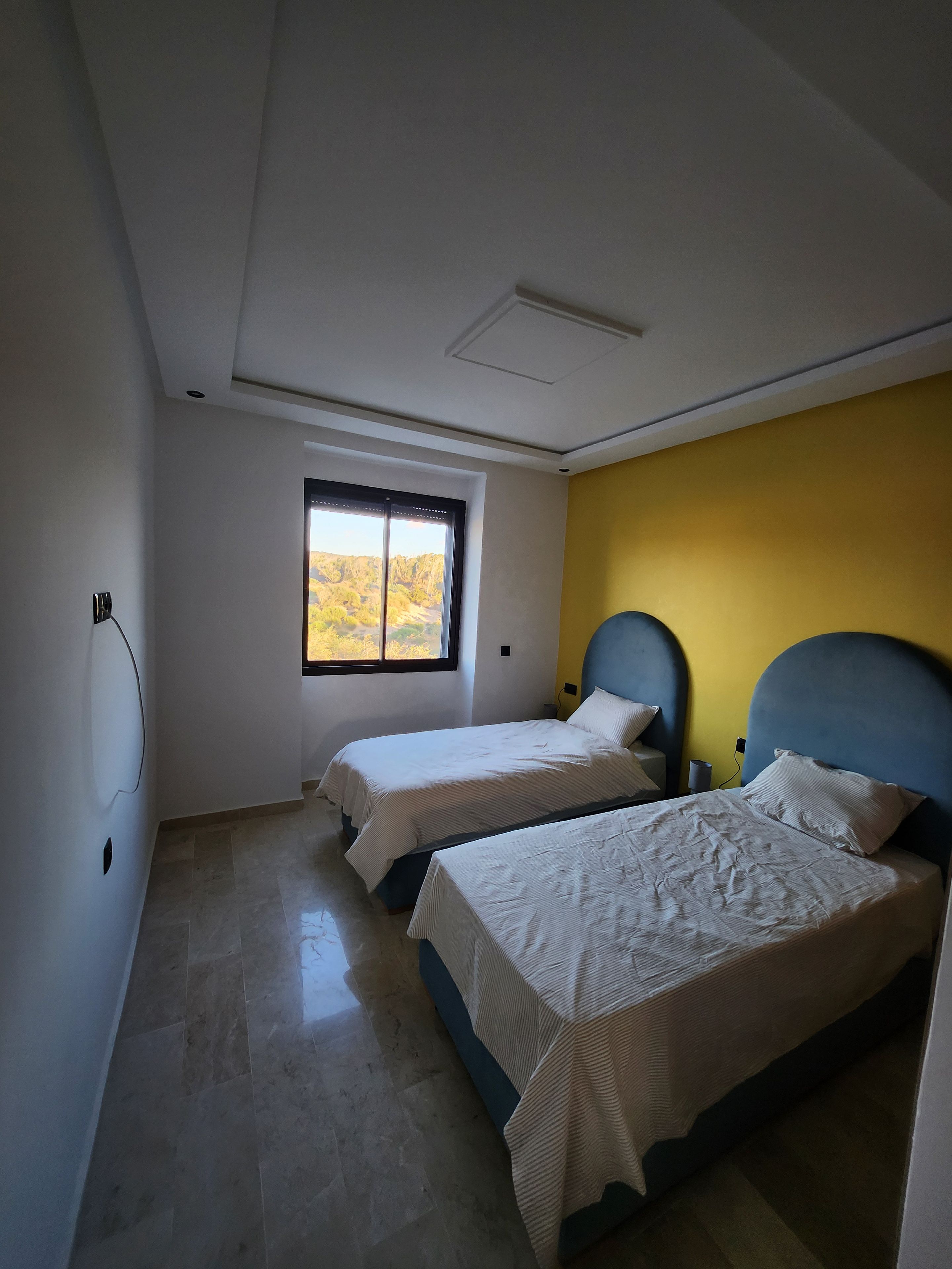 A bedroom with two beds, a window with a view, and a yellow accent wall.
