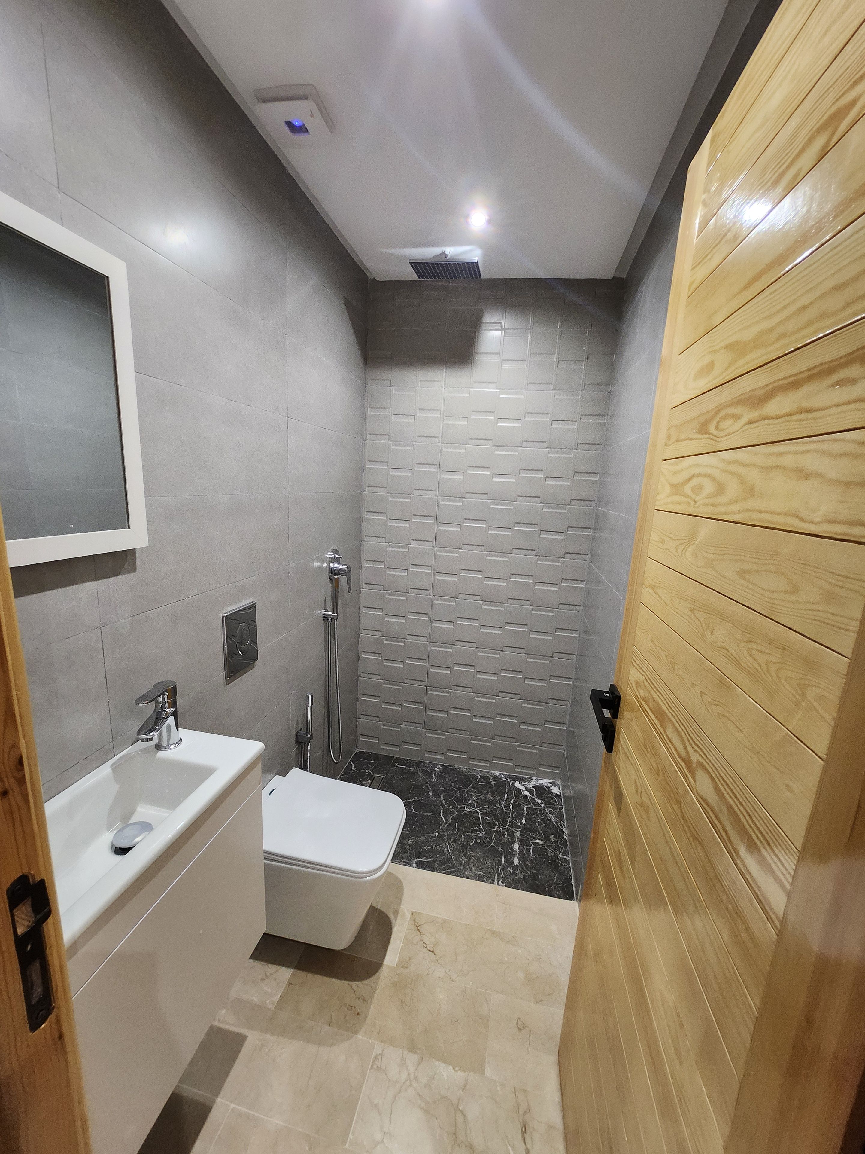 Un bagno con un lavandino bianco, uno specchio, una doccia con soffione manuale e una porta in legno.
