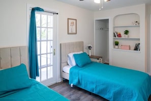 A bedroom with two beds, a window with curtains, a bedside table with a lamp, and a shelf with books and a plant. - The Italian Farmhouse (Cleveland Heights)