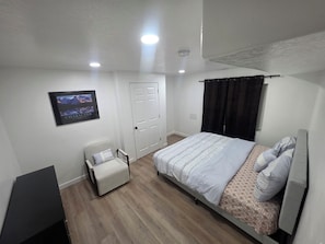 A bedroom with a bed, a chair, a door, and a wall-mounted TV. - Cozy Home Away From Home |Inviting 2 Bedroom! (Saratoga Springs)