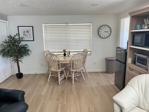 A dining area with a wooden table and chairs, a potted plant, a clock on the wall, and a kitchen with a refrigerator and microwave. - "The Yellowbelle" Pinecraft N. - Sarasota 2b/1b Private Apartment w/ Shared Wall (Sarasota)