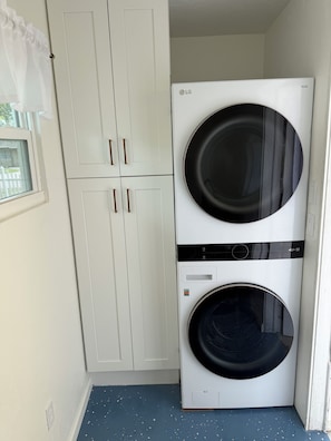 A laundry room with a white washing machine and dryer stacked vertically, a white wardrobe with two doors, and a window with a white curtain. - "The Yellowbelle" Pinecraft N. - Sarasota 2b/1b Private Apartment w/ Shared Wall (Sarasota)