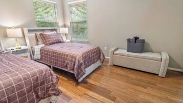 A bedroom with two beds, a nightstand, a lamp, a bench with storage, and a window with blinds.