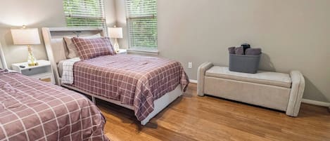 A bedroom with two beds, a nightstand, a lamp, a bench with storage, and a window with blinds.