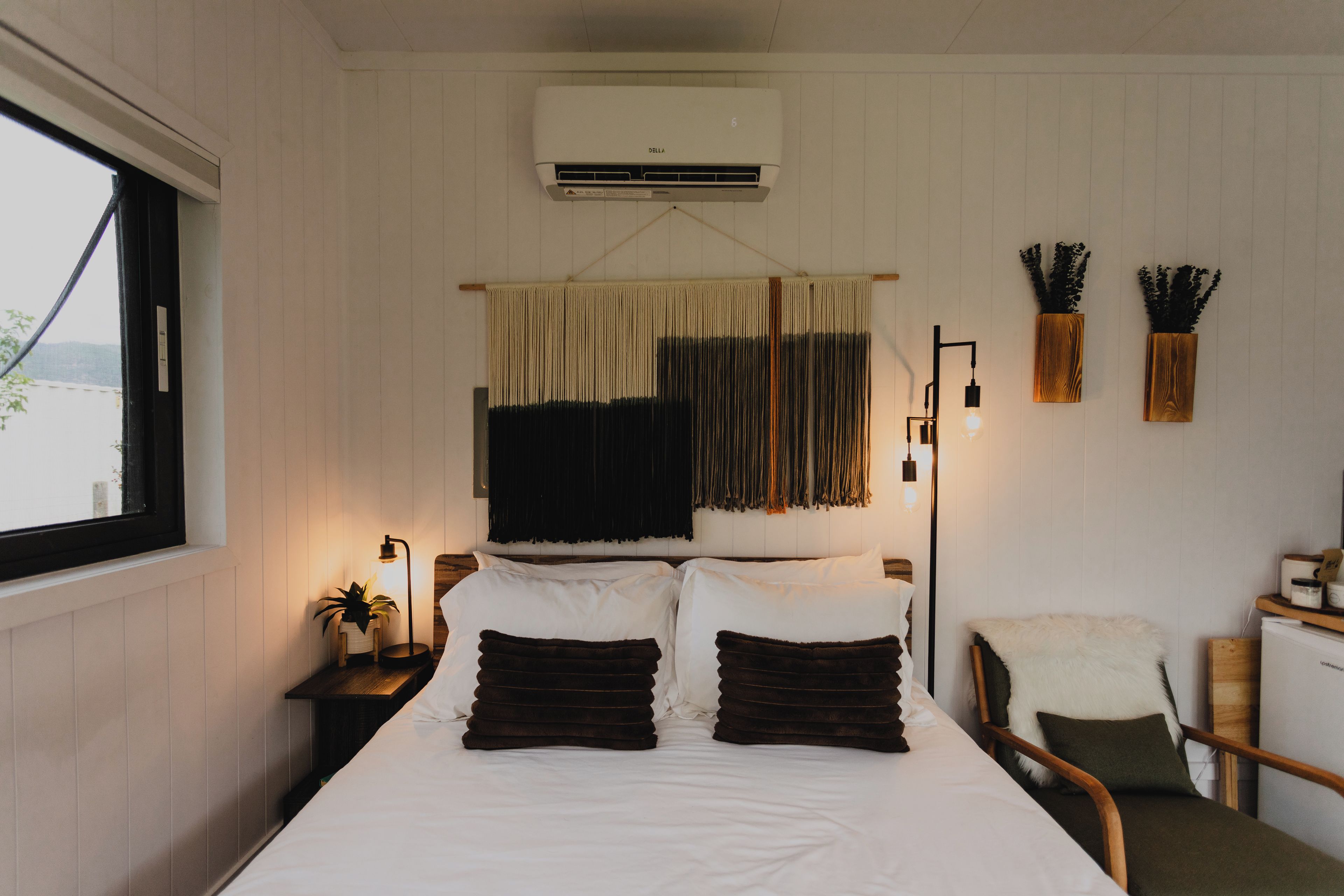 A bedroom with a bed, bedside lamp, air conditioning unit, window, and a chair.
