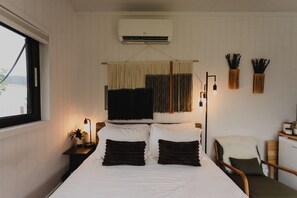 A bedroom with a bed, bedside lamp, air conditioning unit, window, and a chair.