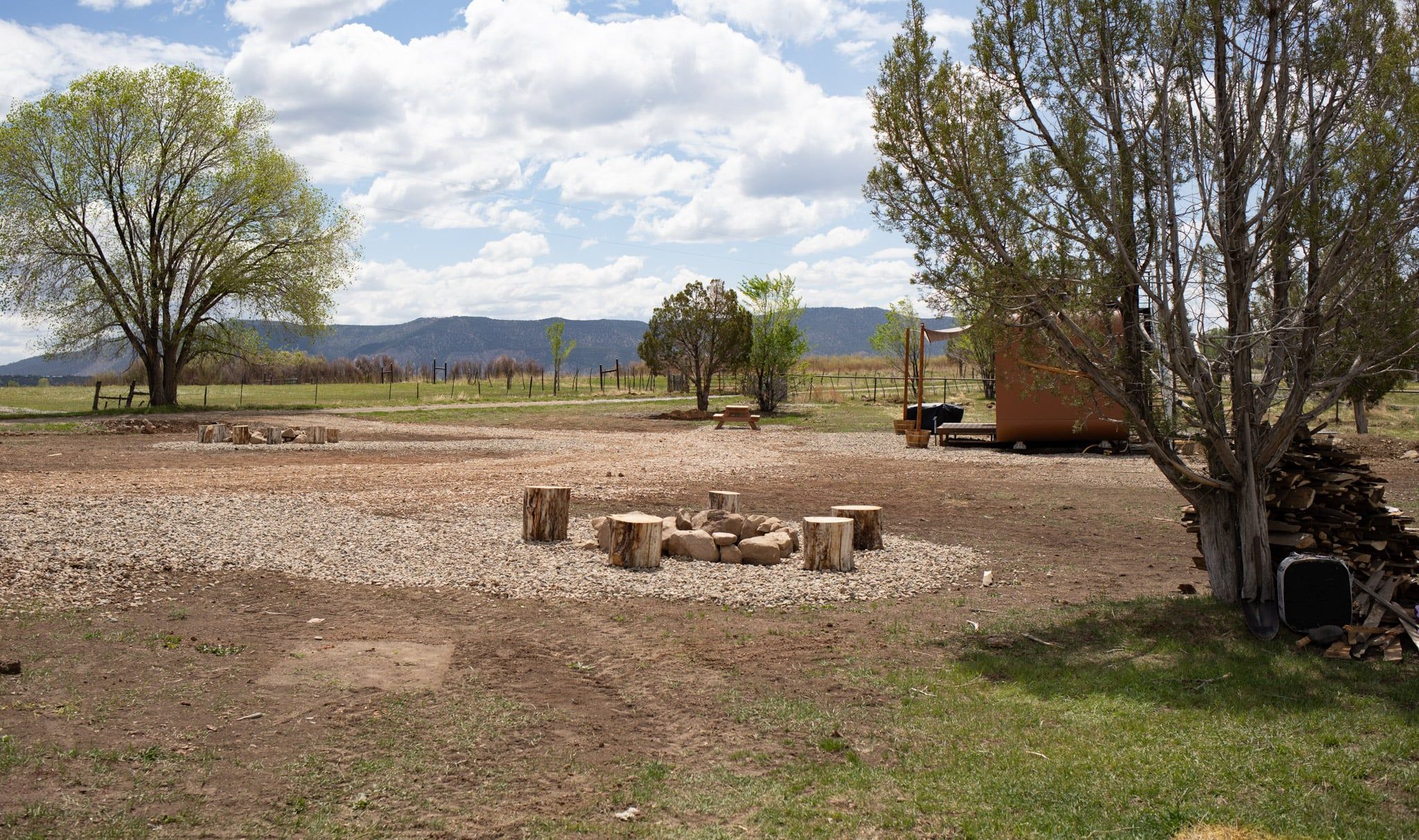 A rural outdoor setting with a dirt ground, scattered wooden stumps, a picnic table, and a pile of firewood.