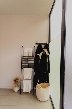 A bathroom with a towel rack holding multiple towels and a black robe. There is a woven basket on the floor and a vase with dried flowers.