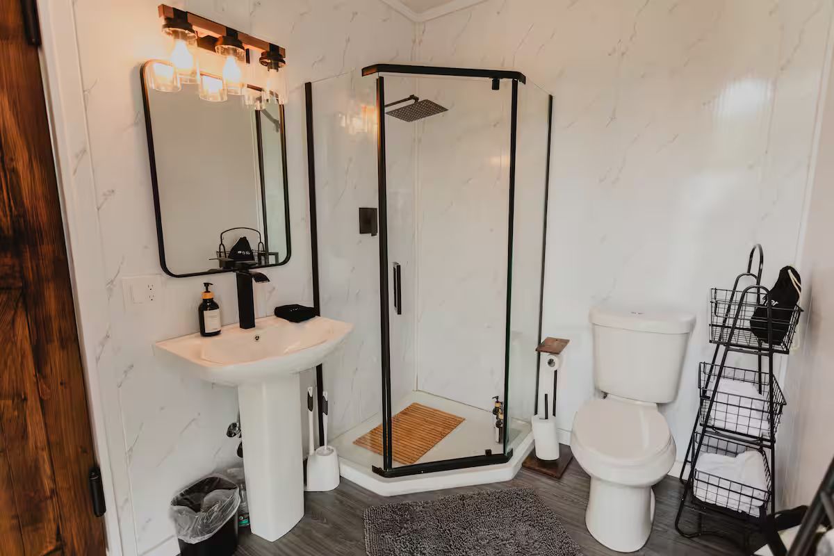 Et badeværelse med en glasafskærmet brusekabine, en hvid vask, et toilet og et spejl med lys.