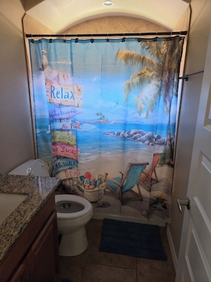 A bathroom with a beach-themed shower curtain, a toilet, a sink, and a chair. - Caravan House RV garage for short term or winter visits (Lake Havasu City)