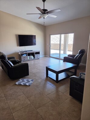 A living room with a ceiling fan, a flat-screen TV, a sofa, a coffee table, and a sliding glass door leading to an outdoor area. - Caravan House RV garage for short term or winter visits (Lake Havasu City)