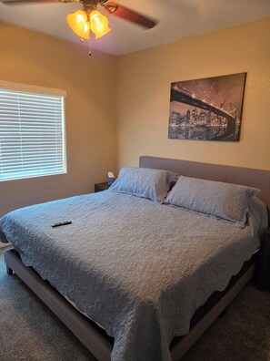 A bedroom with a bed, a nightstand, a ceiling fan, a window with blinds, and a cityscape painting on the wall. - Caravan House RV garage for short term or winter visits (Lake Havasu City)