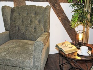 Een knus ingerichte kamer met een groen gestoffeerde fauteuil, een klein rond tafeltje met een boek, glazen, een kopje en een kaars.