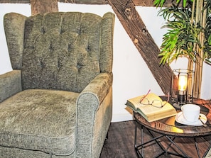 Un rincón acogedor para leer, con un sillón mullido, una mesita redonda con un libro, gafas y una taza, y una planta junto a una viga de madera.