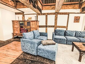 Ein Zimmer mit freiliegenden Holzbalken, einem blau gepolsterten Sofa, einem Holzschrank mit Büchern und einem gemusterten Teppich.