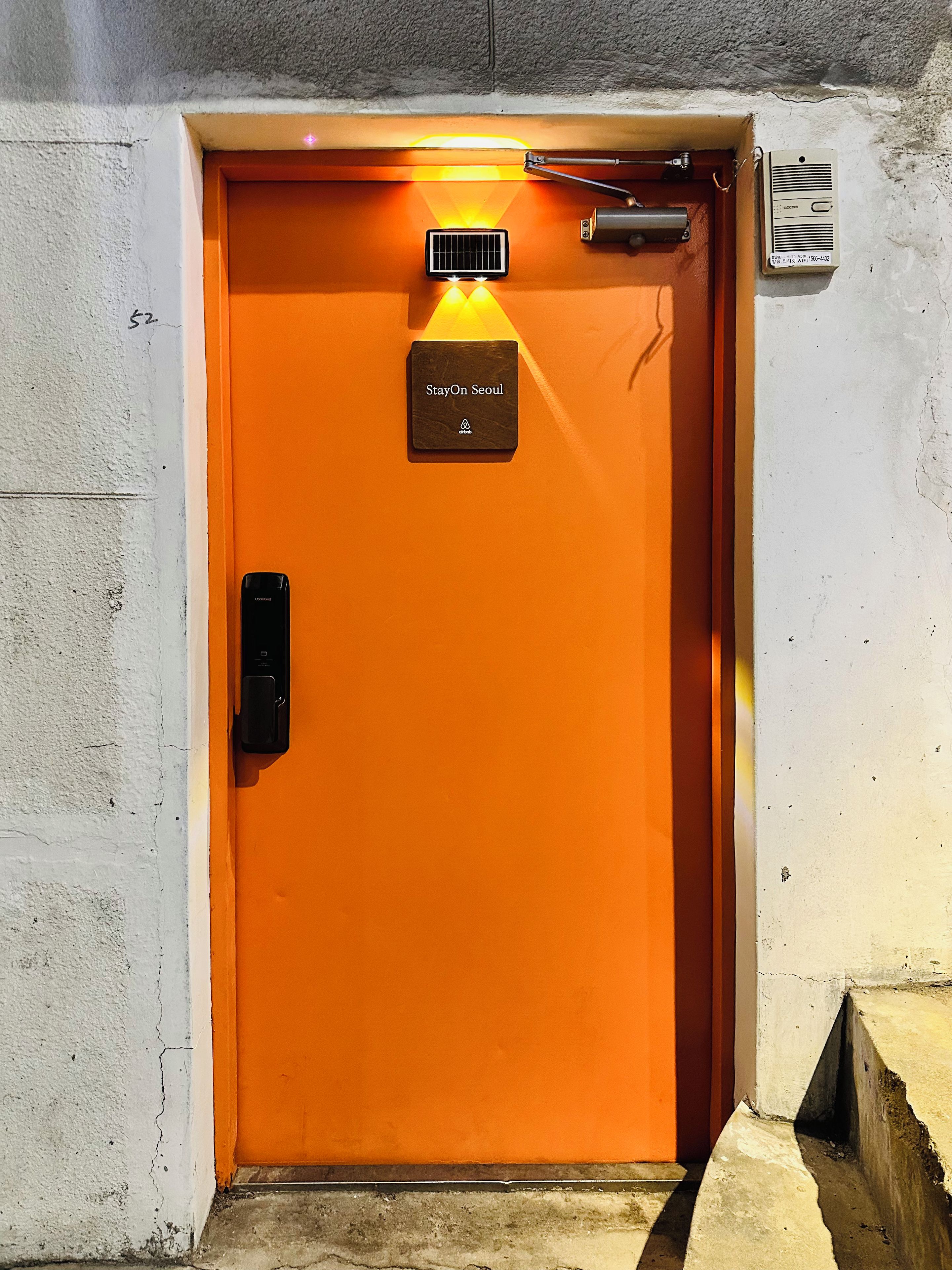 An orange elevator door with a 'StayOn Seoul' sign, a solar panel, and a security device.