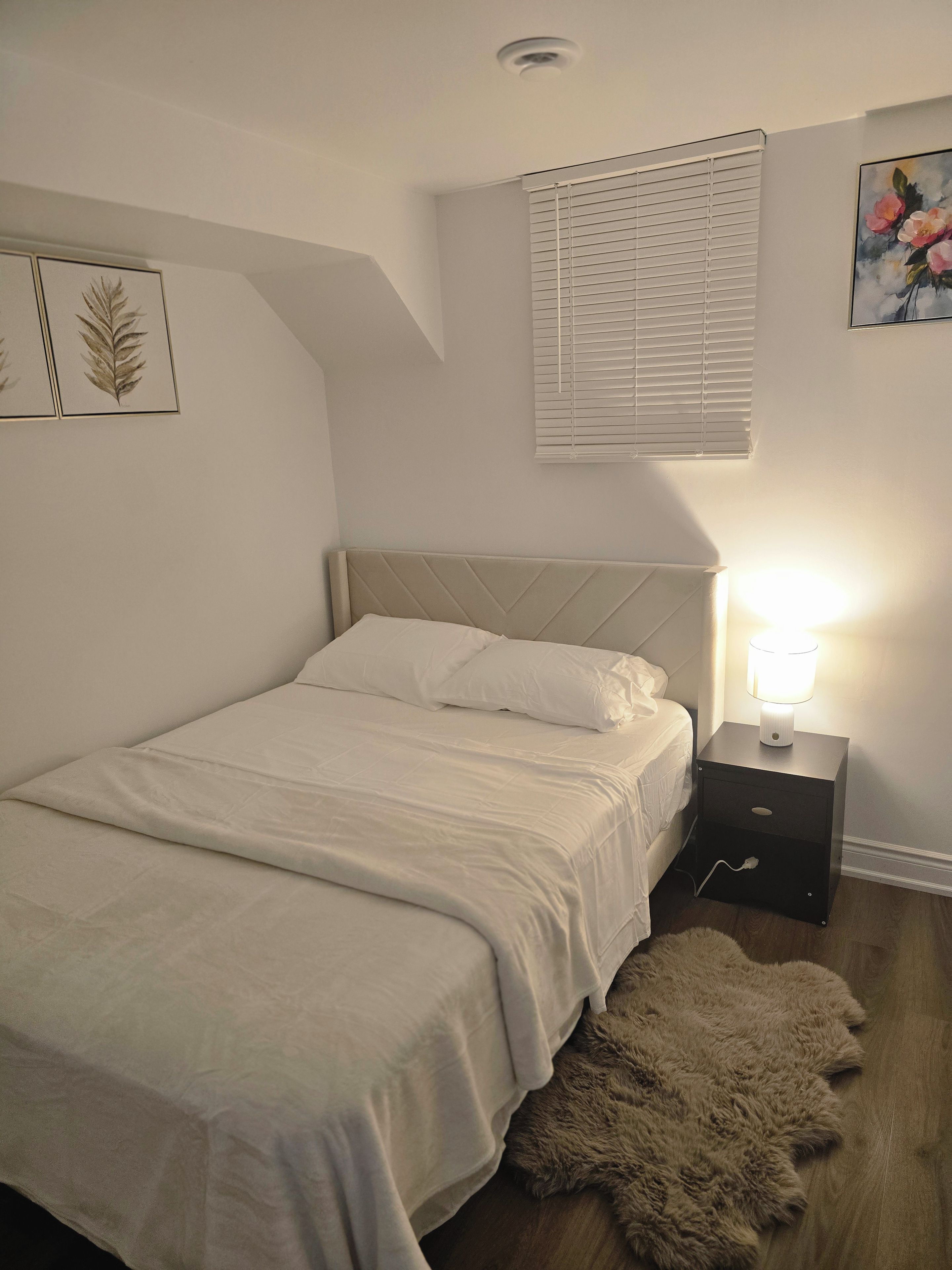 Ein Schlafzimmer mit einem Bett, einem Nachttisch mit Lampe, einem Fenster mit Jalousien und einer Wand mit gerahmten Kunstwerken.