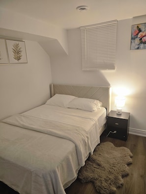 Ein Schlafzimmer mit einem Bett, einem Nachttisch mit Lampe, einem Fenster mit Jalousien und einer Wand mit gerahmten Kunstwerken.