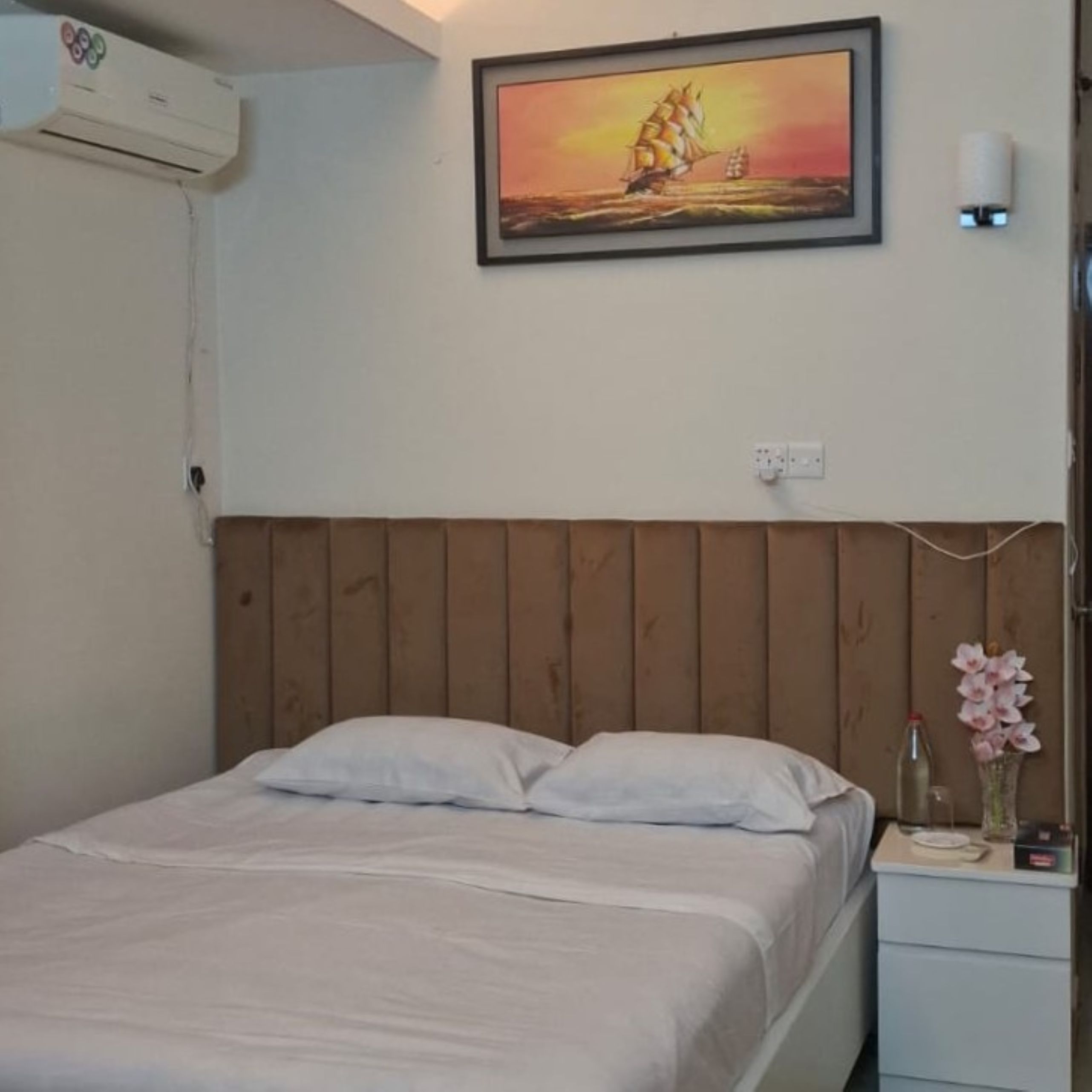 A bedroom with a wooden headboard, a bed with white bedding, a nightstand with a vase of flowers, and a painting of a sailboat on the wall.
