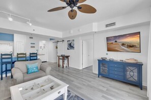 A spacious living room with a ceiling fan, a dining area with chairs, a kitchen with a white countertop, and a large painting on the wall. - Shores of Panama 2BD Condo| Beach, Pools & Gulf Views (Panama City Beach)