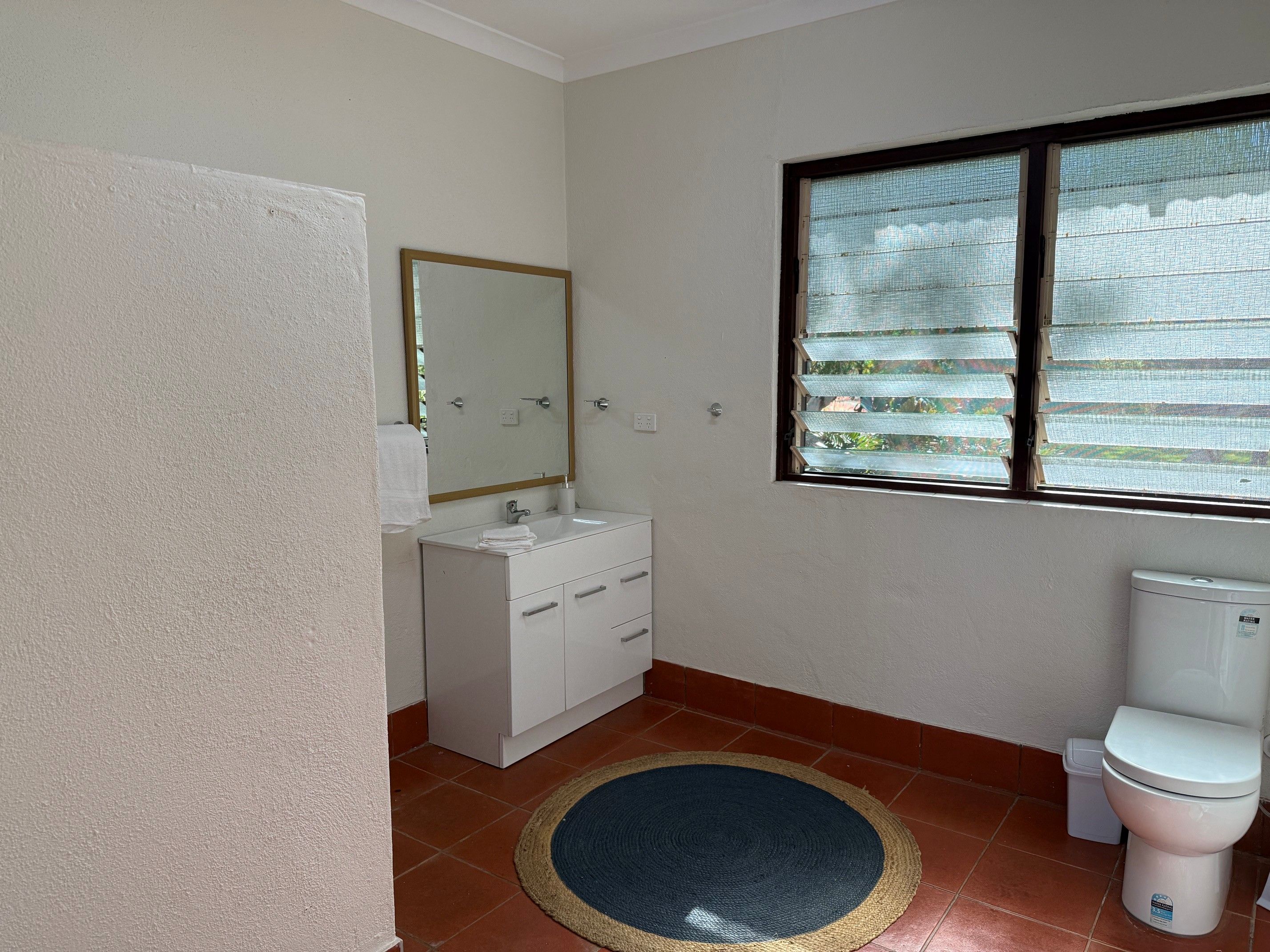 Un baño con lavamanos blanco, un espejo, inodoro y ventana con persianas.