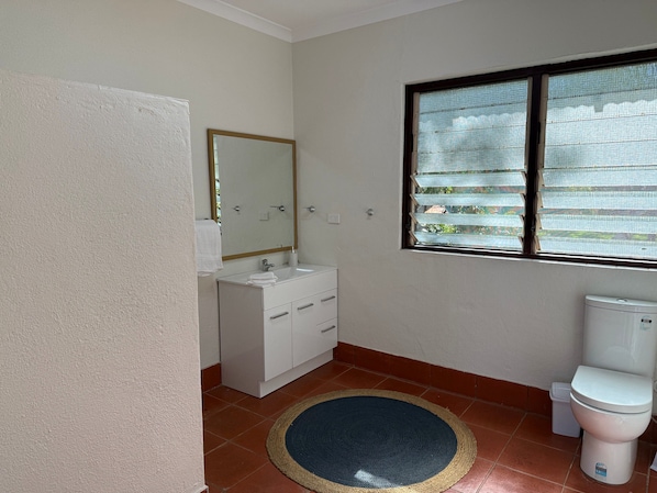 Un baño con lavamanos blanco, un espejo, inodoro y ventana con persianas.