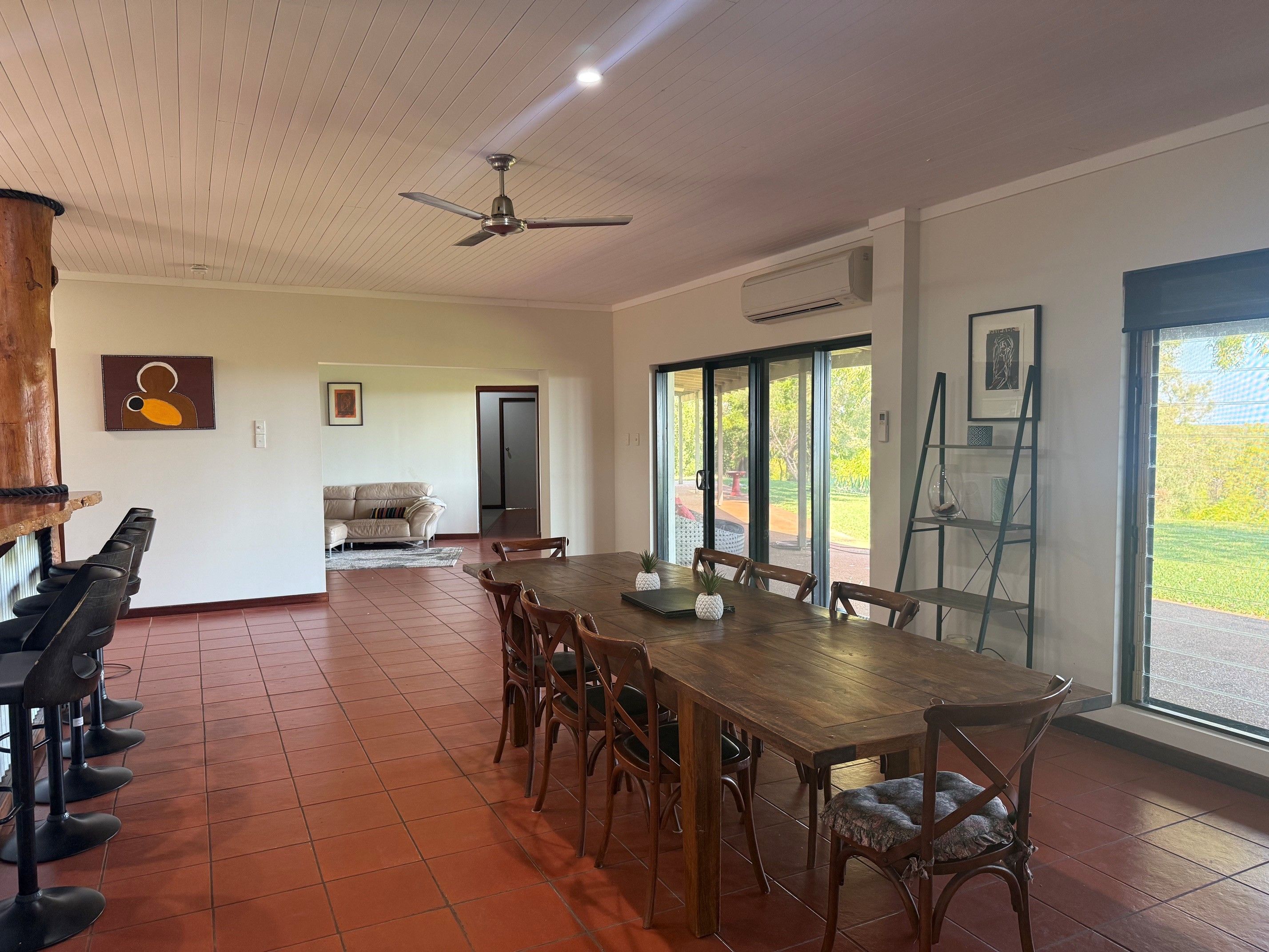 A spacious dining area with a long wooden table and chairs, a ceiling fan, and a large window with a view of the outdoors.