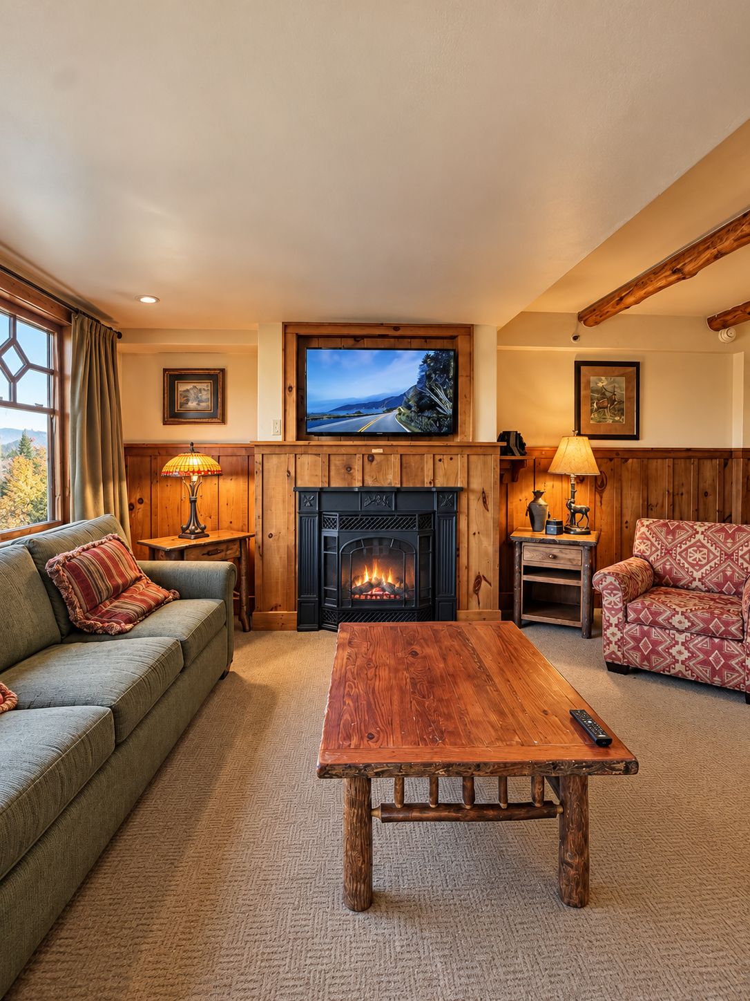 A cozy living room with a fireplace, a television mounted above it, a sofa, a patterned armchair, and a wooden coffee table.