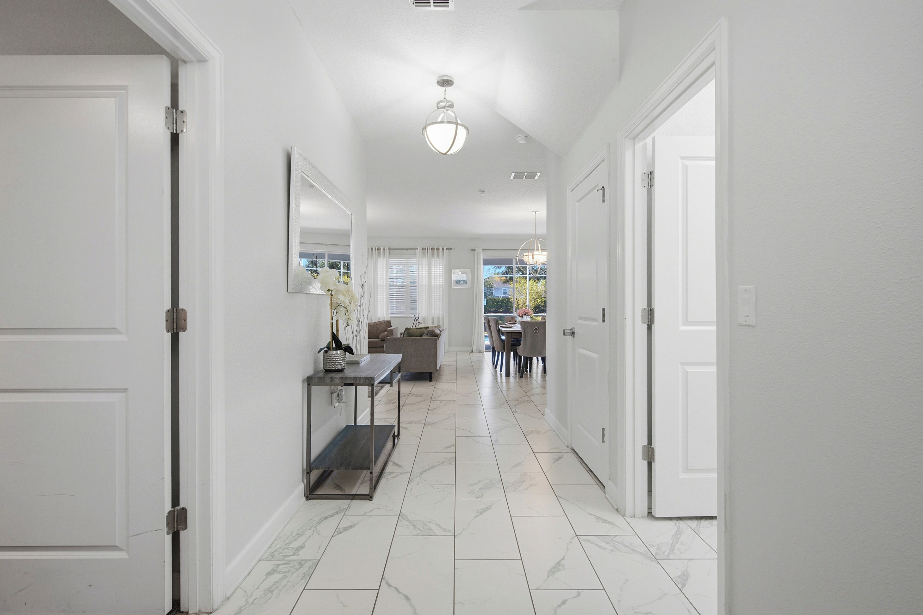 A hallway with white walls, a marble floor, and doors leading to other rooms. There is a table with a vase and a chair, and a dining area with a table and chairs in the background.