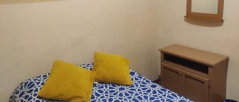A room with a bed, two yellow pillows, a wooden cabinet, a painting, and a mirror on the wall.