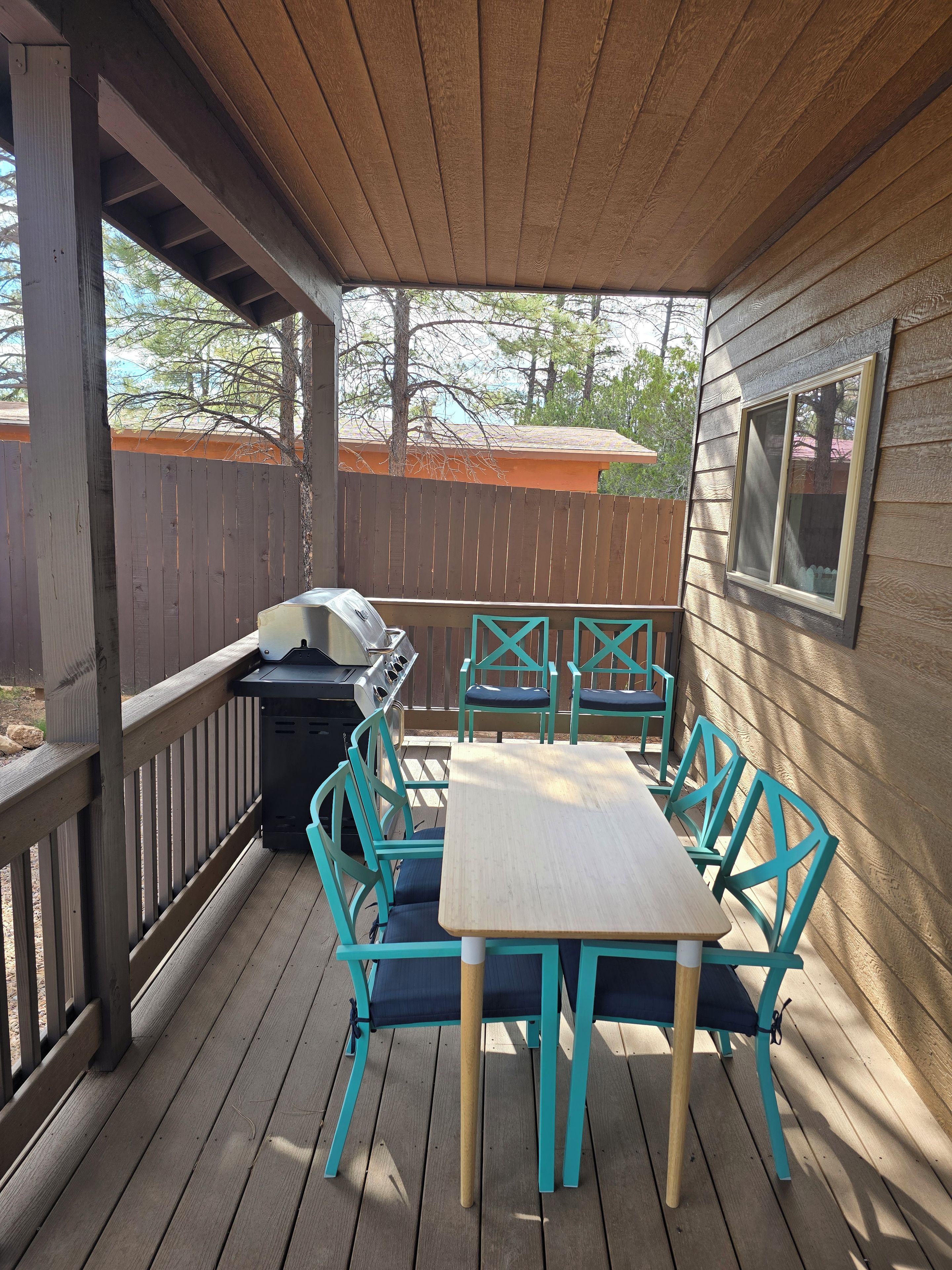 A covered outdoor patio with a wooden table and chairs, a grill, and a view of a neighboring house.