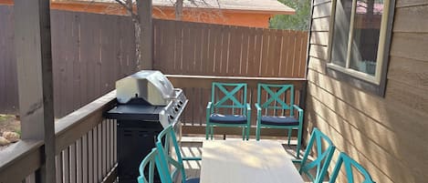 A covered outdoor patio with a wooden table and chairs, a grill, and a view of a neighboring house.
