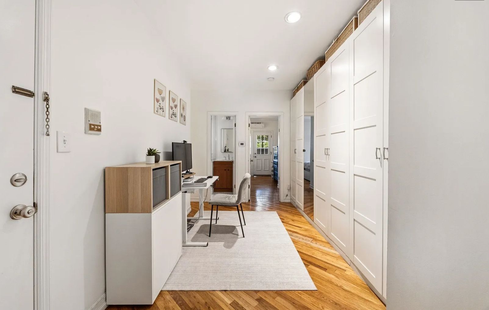 A narrow hallway with a desk, chair, and a television on the left. There are white doors on both sides, and a wooden floor.
