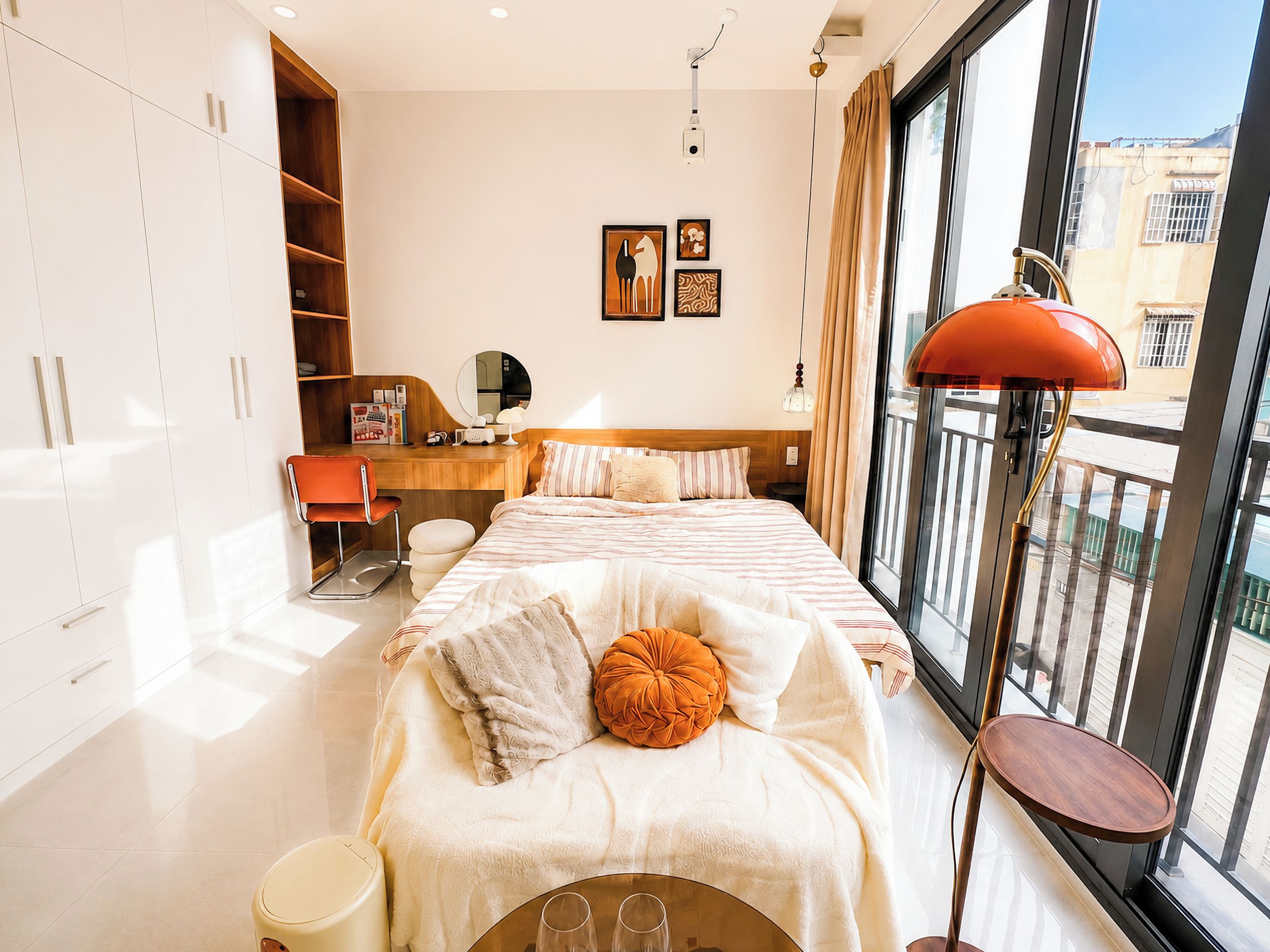 A modern bedroom with a bed, a desk, and a chair. There is a window with a view of buildings outside.
