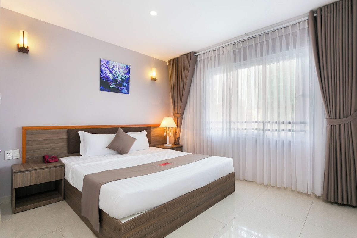 A hotel room with a large bed, a bedside table with a lamp, a wall-mounted light, and a window with curtains.