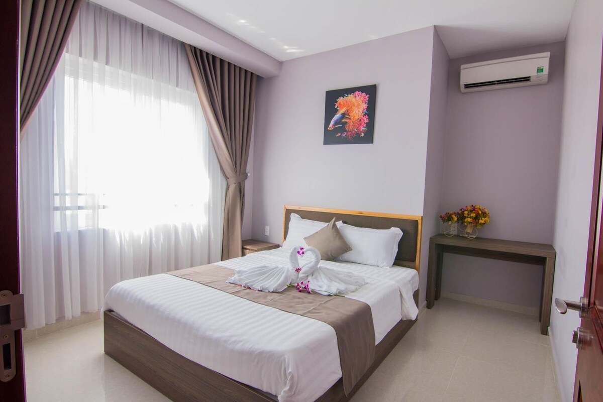 A hotel room with a bed, a nightstand, an air conditioning unit, and a window with curtains.