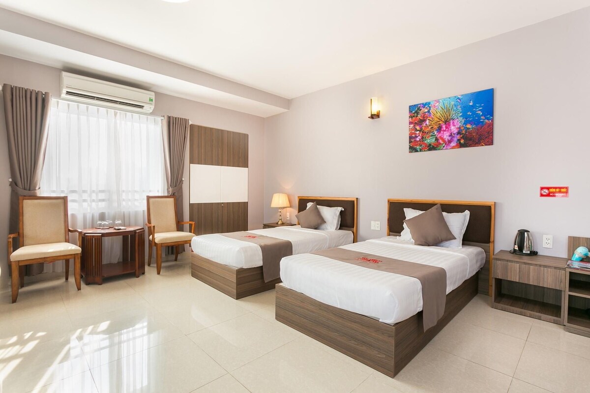 A hotel room with two beds, a desk, chairs, an air conditioning unit, a painting, and a coffee machine.