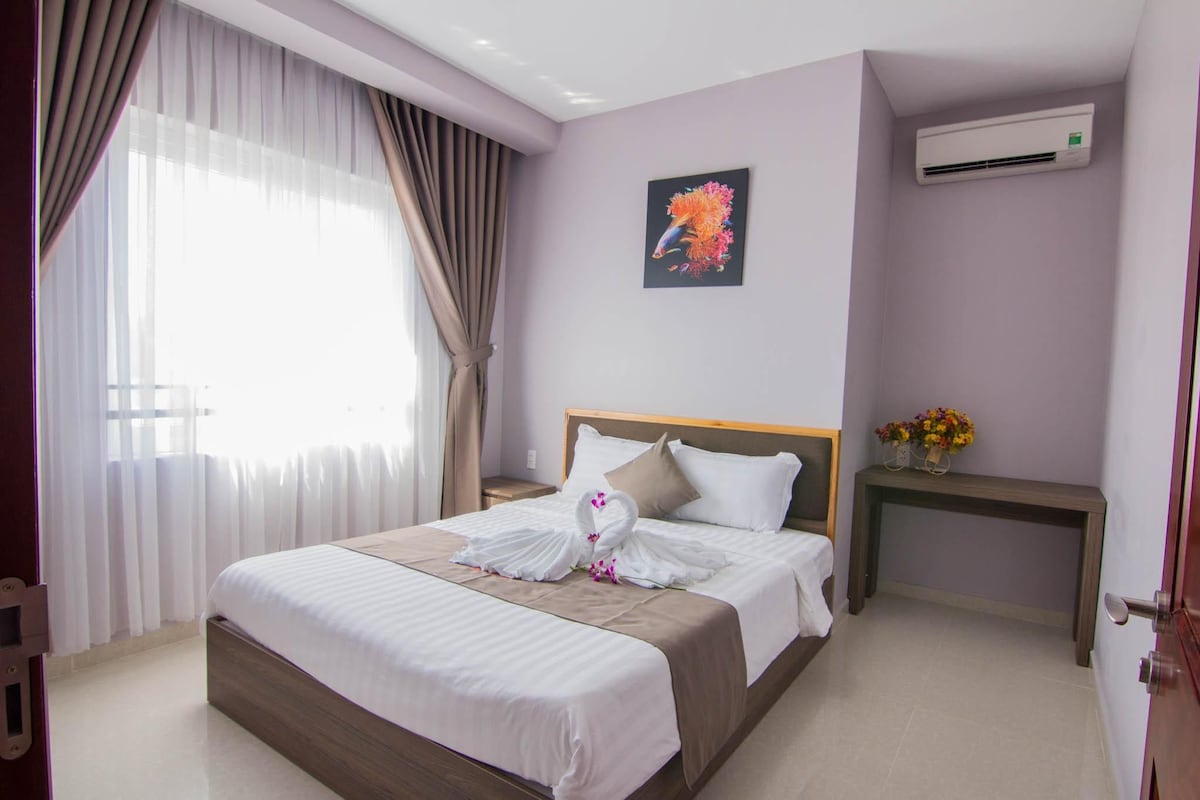 A hotel room with a bed, a nightstand, an air conditioning unit, and a window with curtains.