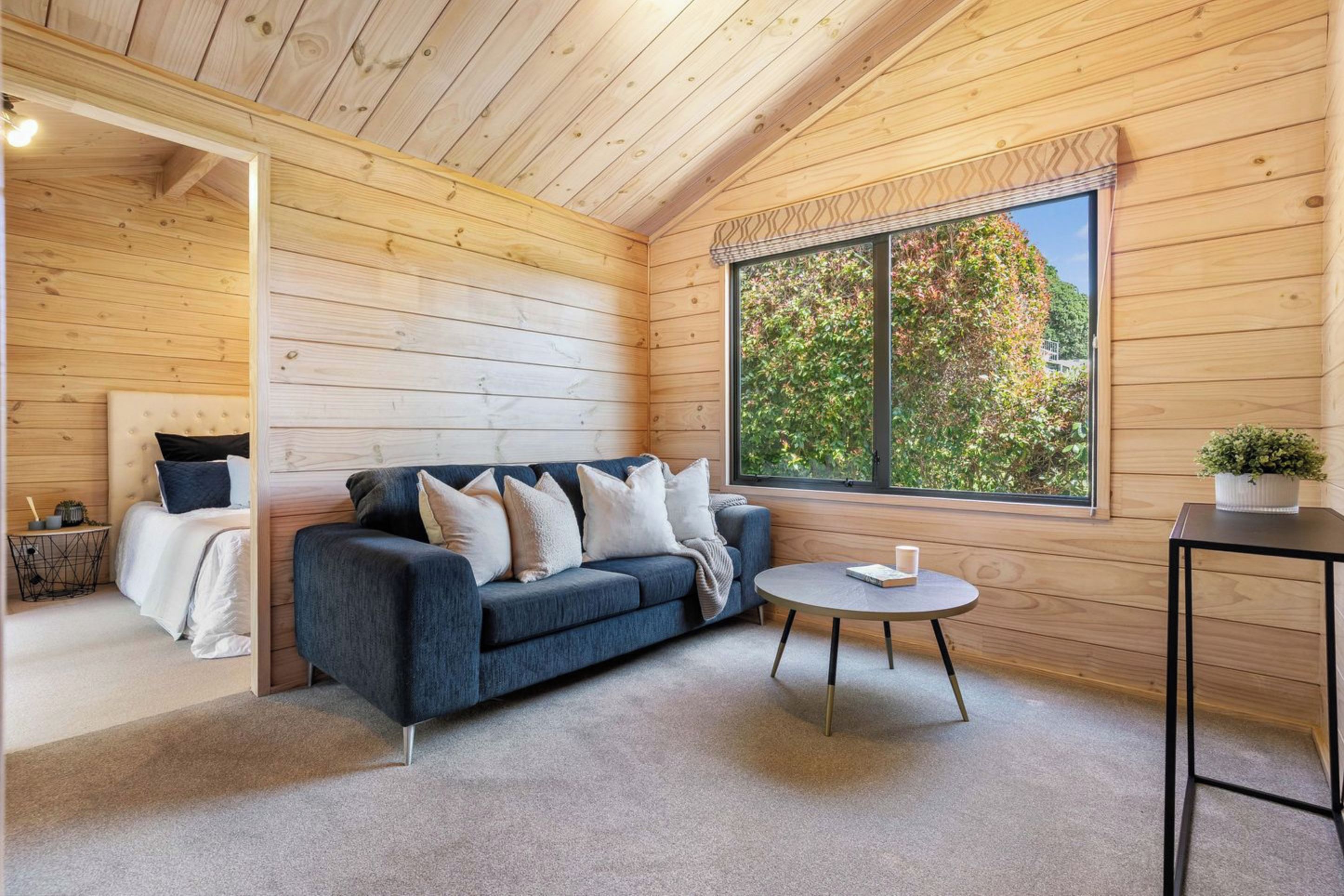 A cozy room with a blue sofa, a round table, and a large window with a view of greenery. - Family coastal retreat, 4min walk to the Beach (Pauanui)