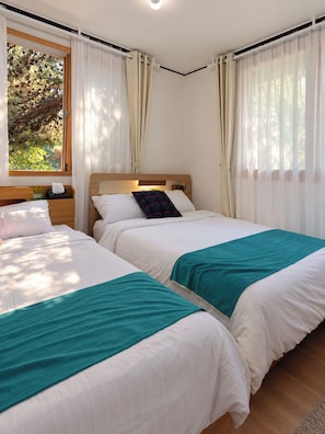 Ein Doppelzimmer mit zwei Betten, einem Holz-Kopfteil, einem Fenster mit Vorhängen und Blick auf Bäume.