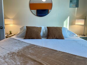 A neatly made bed with a white comforter and two brown pillows, a round mirror on the wall, and two bedside lamps. - Heart of Tobin Hill! Walk to The Pearl, River Walk! King Bed, Pac-Man, Sleeps 4 (San Antonio)