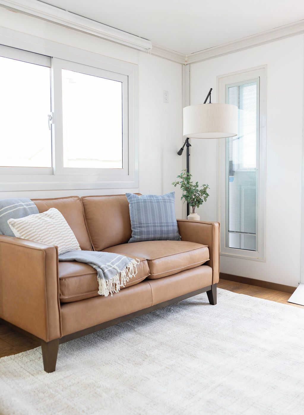 A tan leather sofa with cushions, a white rug, and a floor lamp in a bright room with large windows.