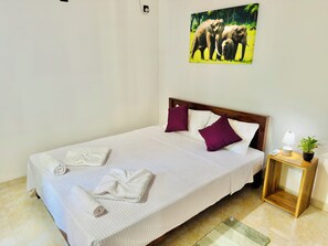 Cama bem arrumada com roupa de cama branca e duas almofadas roxas, uma pequena mesa de cabeceira com luminária e uma planta, e um quadro na parede com elefantes.
