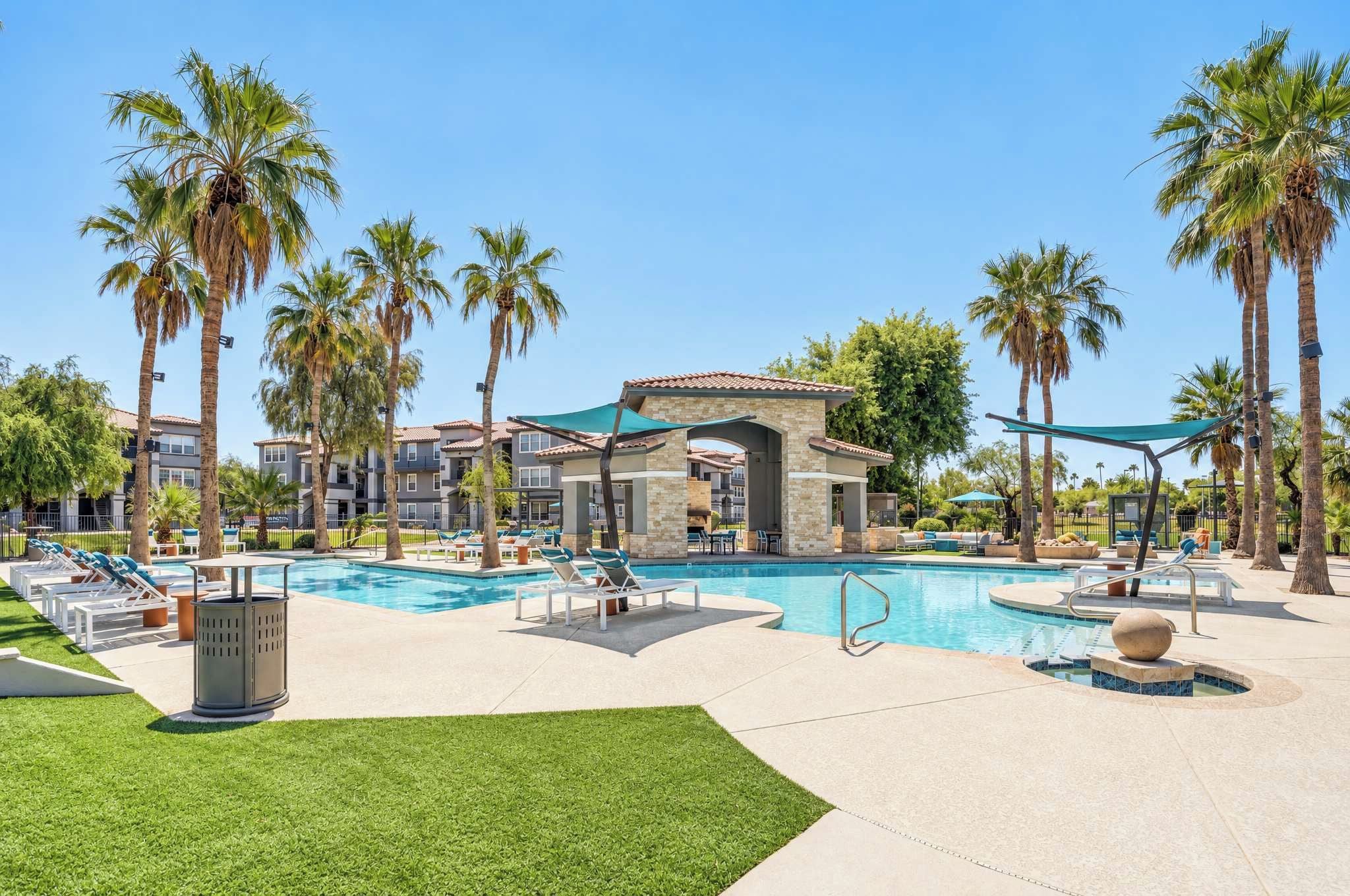 A resort with a swimming pool, lounge chairs, palm trees, and a building with a tiled roof.