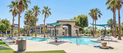 A resort with a swimming pool, lounge chairs, palm trees, and a building with a tiled roof.