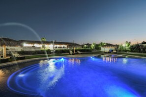 Ein Schwimmbad bei Nacht mit blauer Beleuchtung, strohgedeckten Gebäuden und Liegestühlen.