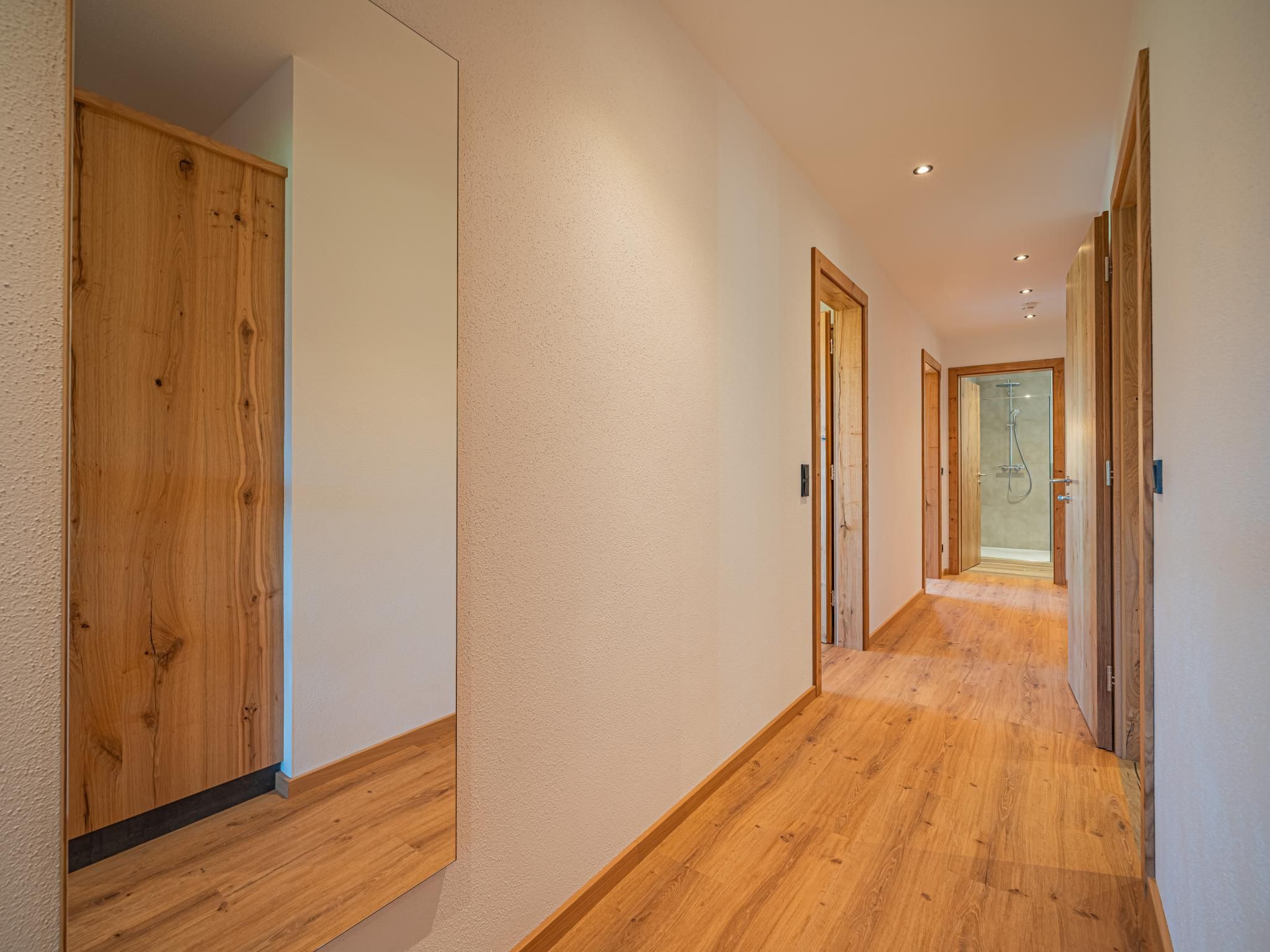 A hallway with wooden flooring and doors, leading to a bathroom with a glass shower.