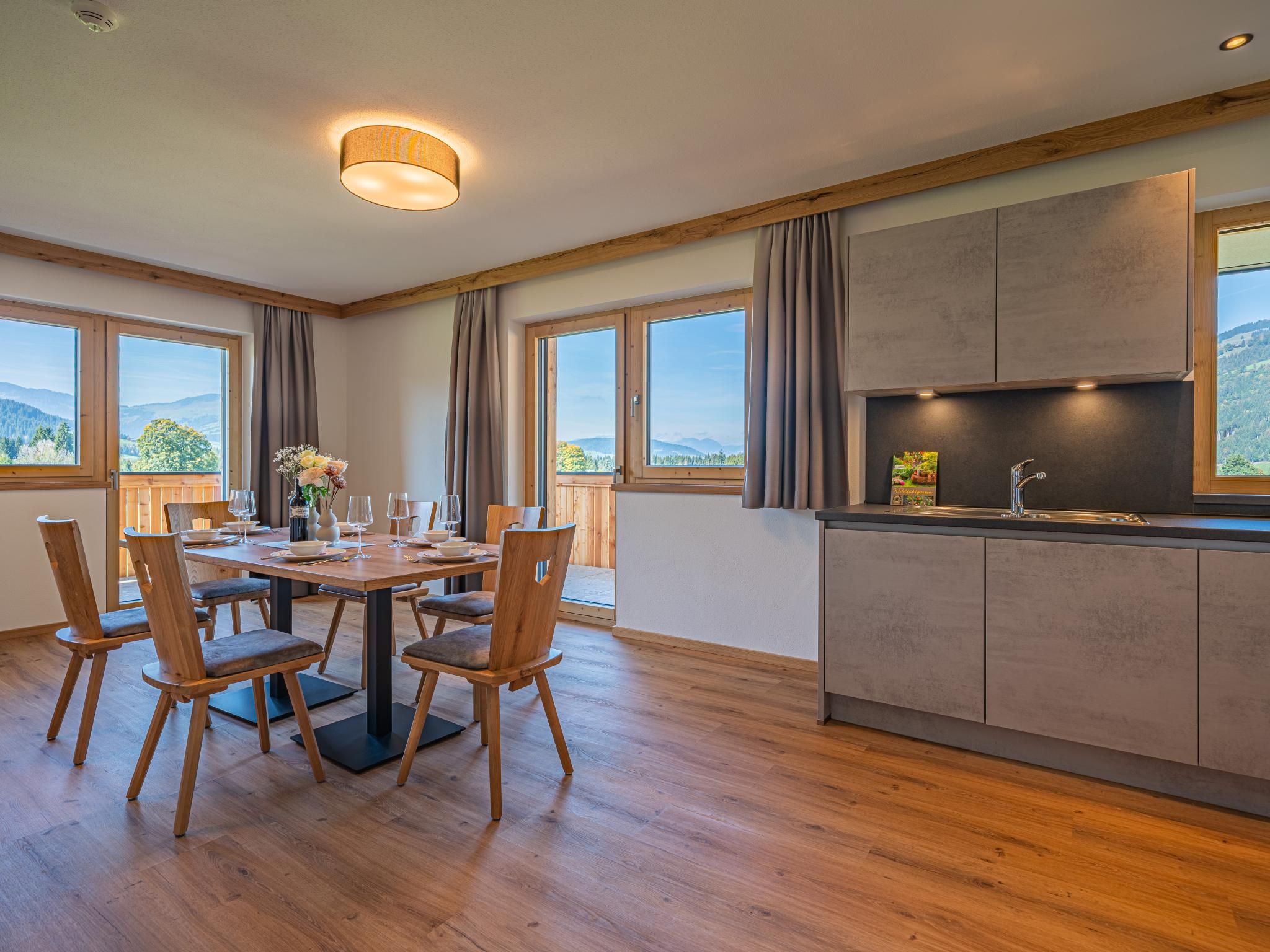Un comedor con mesa y sillas de madera, una cocina con fregadero y armarios, y ventanas amplias con vistas al exterior.
